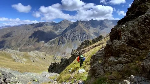 Two hikers are walking through difficult mountainous terrain on a cliff slope Stock Footage 252765910