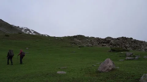 Two hikers traverse grassy hillside with rocky terrain and snow-capped mountains Video stock 340392092