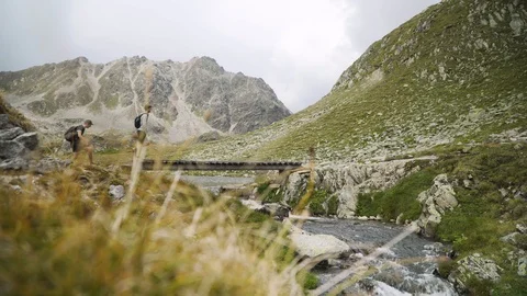 Two hikers walking over bridge along river with mountain panorama, 4K Stock Footage 129611658