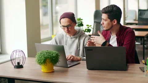 Two hipster coworkers sharing ideas about project sitting with laptops in Stock Footage 219800218