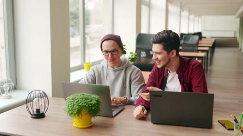 Two hipster coworkers sharing ideas about project sitting with laptops in Stock Footage 219800295