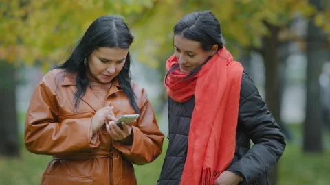 Two hispanic girls stand in autumn park using telephone women ordering food taxi Stock Footage