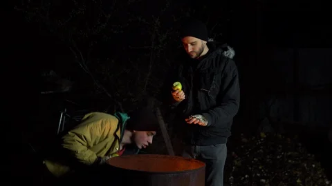 Two homeless young men stand by the fire and eat apples. Men stand at night near Stock Footage 129558595