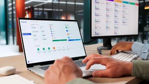 Two HR Managers Discuss Payroll Software Data On Laptops In Office. Stock Photos