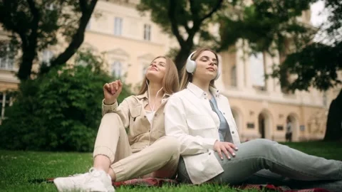 Two incredibly beautiful blonde young women in headphones sitting on the green Stock Footage 197008149