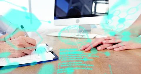 Two individuals at desk, one writing, another using a computer Stockfoto's