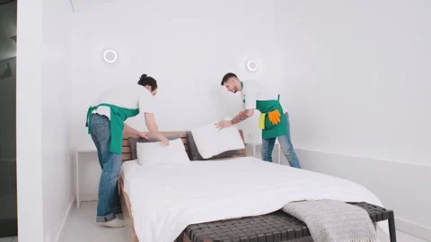 Two individuals expertly arranging a white sheet on a bed in a bright, modern Stock Footage 305277301