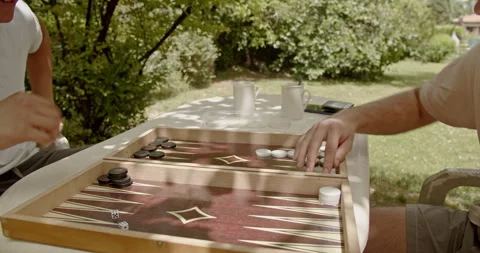 Two individuals playing backgammon on a wooden board table outside. The scene is Stock Footage 282562536
