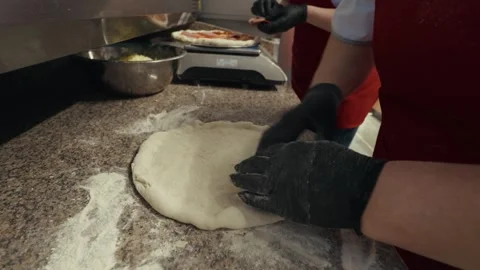 Two individuals in red aprons prepare pizza dough by hand on a countertop, with Vídeo Stock 332736606