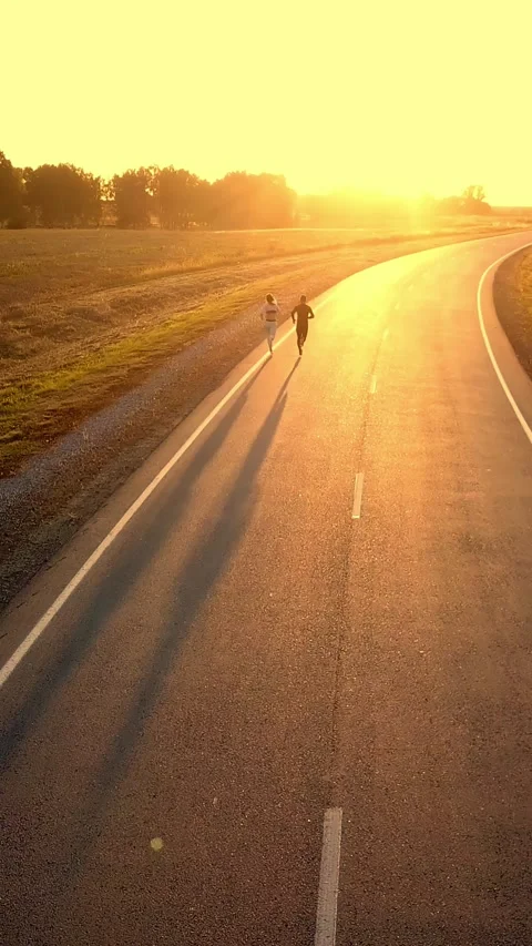 Two individuals walking along a winding road at sunset, casting long shadows in 스톡 동영상 313260642