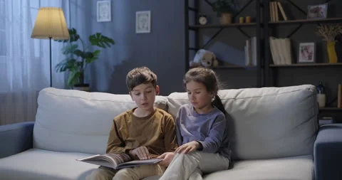Two kids reading book, looking at pictures, turning pages sitting on the sofa in Stock Footage 269850777
