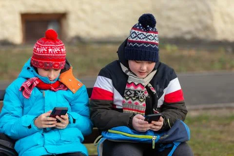 Two kids sit on a bench in winter hats and coats while using their smartphone 写真素材