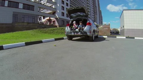 Two kids sit in car boot near dwelling house at spring sunny day Stock Footage 42752355
