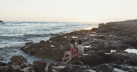Two kids sitting on a rockey beach during sunset Stock Footage 65259768