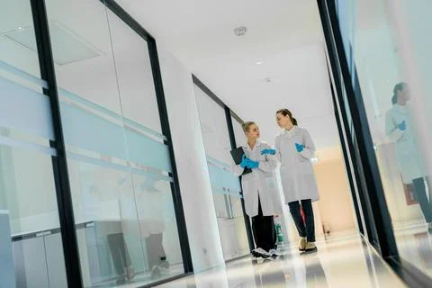 Two lab technicians walk in a hallway discussing work while wearing lab coa.. 写真素材