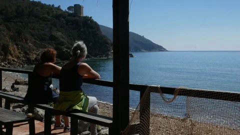 Two Ladies rest seated in front of the sea in a sunny day Stock Footage 105219005