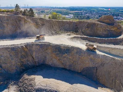 Two large trucks are moving on dirt paths in a quarry. Dust rises as they nav Stock Photos