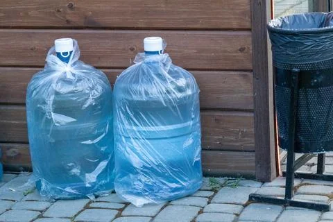 Two Large Water Bottles Wrapped in Plastic Bags Next to a Trash Can in a Ca.. Stock Photos