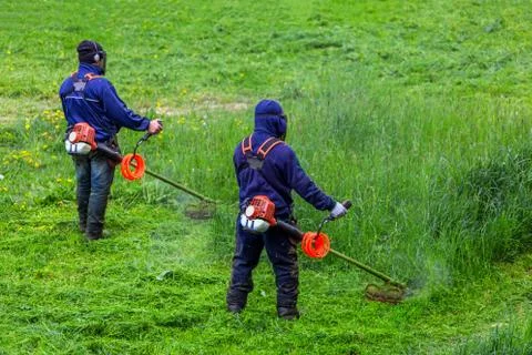 Two lawnmower men with string trimmer and face mask trimmong grass - close-up 库存照片