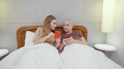 Two lesbians lie in bed before sleep, each using their smartphone and scrolling  Stock Footage 296303074