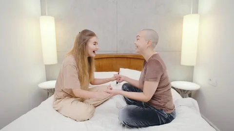 Two lesbians sit on the bed, holding hands and happily chatting, sharing a joyfu Stock Footage 296303155