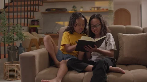 Two little Asian sisters read a book together on a living room sofa Stock Footage 339865742