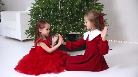 Two little sisters in red dresses play near Christmas tree indoors having fun  Stock Footage 295417232