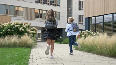 Two little students, a boy and a girl, run towards the school building. The Stock Footage 212890343