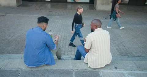 Two male friends sitting on steps on break handling food box, sipping drink Vidéo 343099476