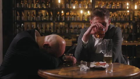 Two men are sitting at a bar table, looking distressed and tired. One man has Stock Footage 289111610