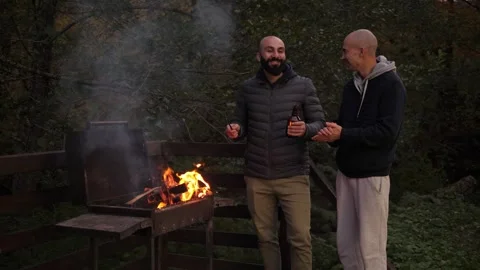 Two men are standing around a campfire in the evening, talking and drinking beer Stock Footage 294678872