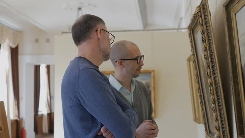 Two men in an art gallery look at a painting and talk about art Stock Footage 128477978