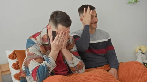 Two men on bed, one covering his face with hands while holding a phone and .. 스톡 동영상 339217414