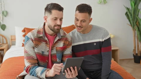 Two men on bed, one points finger to tablet while both lean in viewing the .. Stock Footage 339217642