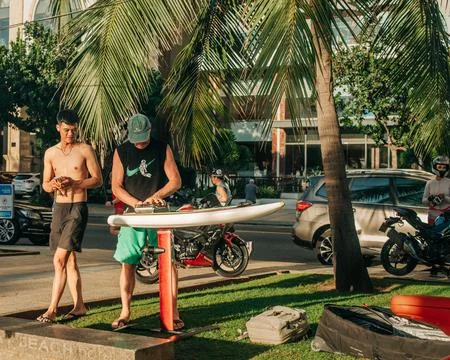 Two men carrying and preparing a paddleboard under palm trees beside a busy Stock Photos