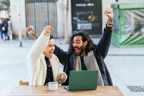 Two men celebrating while using a laptop outdoors at a cafe. Stock Photos