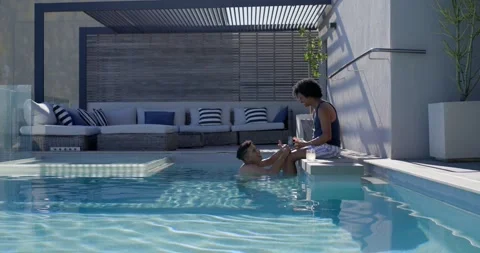 Two men chatting poolside, man leaning in water lifting legs onto man reaching Stock Footage 339652218