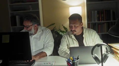Two men at a desk in a building, one typing on a laptop while the other rea.. Stock Footage 339214322