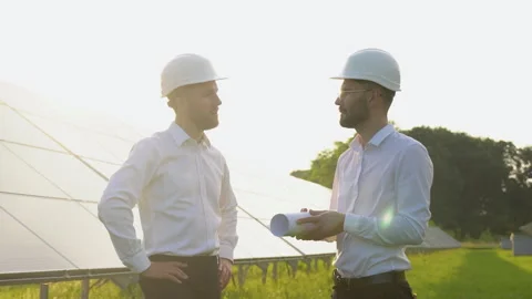 Two men in hard helmets discussing project at solar farm. Male engineers with Vidéo 245575813