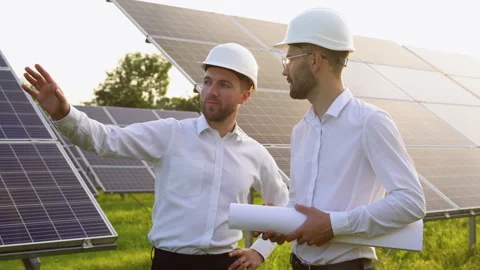 Two men in hard helmets discussing project at solar farm. Male engineers with Vidéo 245577372