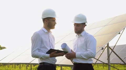 Two men in hard helmets discussing project at solar farm. Male engineers with Vidéo 245578149