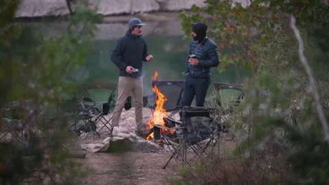 Two men holding coffee cups talking around morning campfire in front of Stock Footage