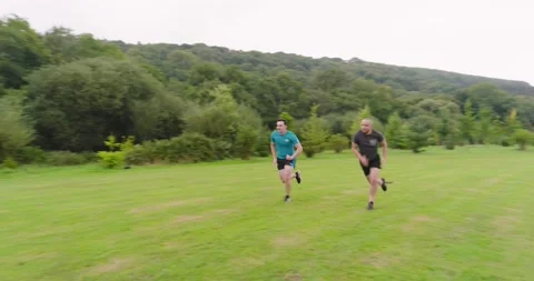 Two men jogging on grassy field surrounded by lush green trees Stock Footage 345571383