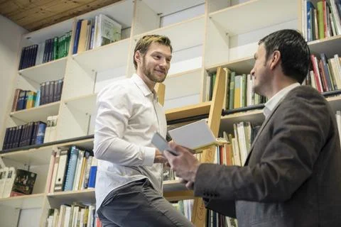 Two men looking at each other in front of bookshelf, Bavaria, Germany Stock Photos