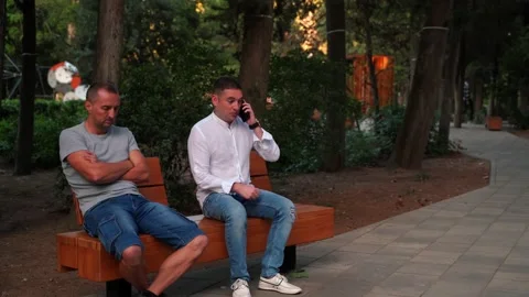 Two men in the park on a bench. One man is asleep, the other is on the phone. Stock Footage 139593496
