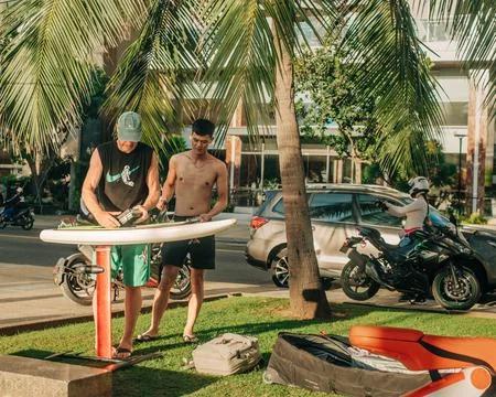 Two men preparing a paddleboard beside a tropical seaside promenade with palm Stock-Fotos