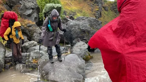 Two men in raincoats help a young woman to jump over a stormy river Stock Footage 140895351