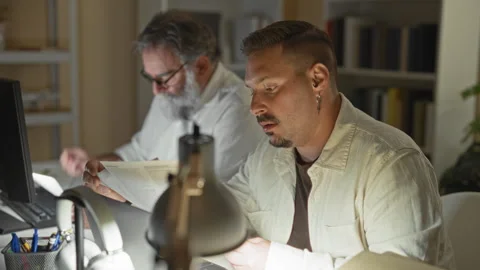 Two men read documents at a desk, one holds glasses and the other rubs his .. Stock Footage 339215580