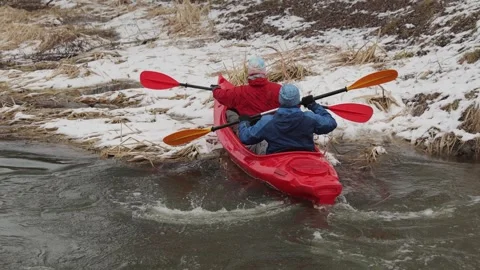 Two men in a red kayak are swimming to a snow-covered shore with reeds. Video stock 299710757