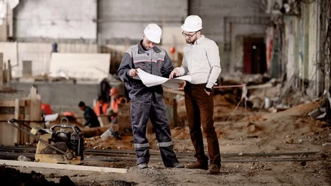 Two men in safety helmets standing in construction area and looking at scheme of Stock Footage 77626677